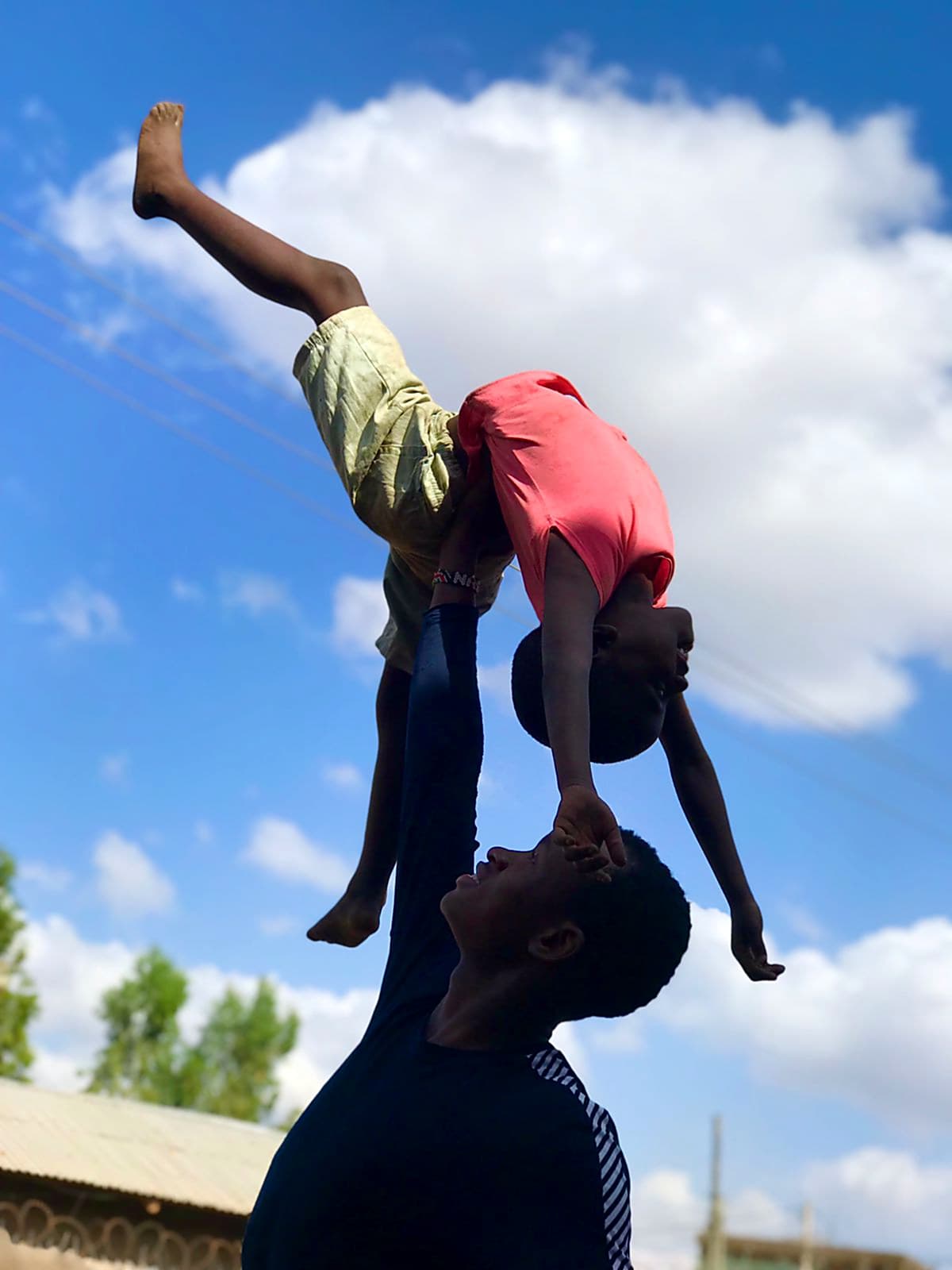 A Nafsi acrobat lifting a child overhead against the Nairobi sky